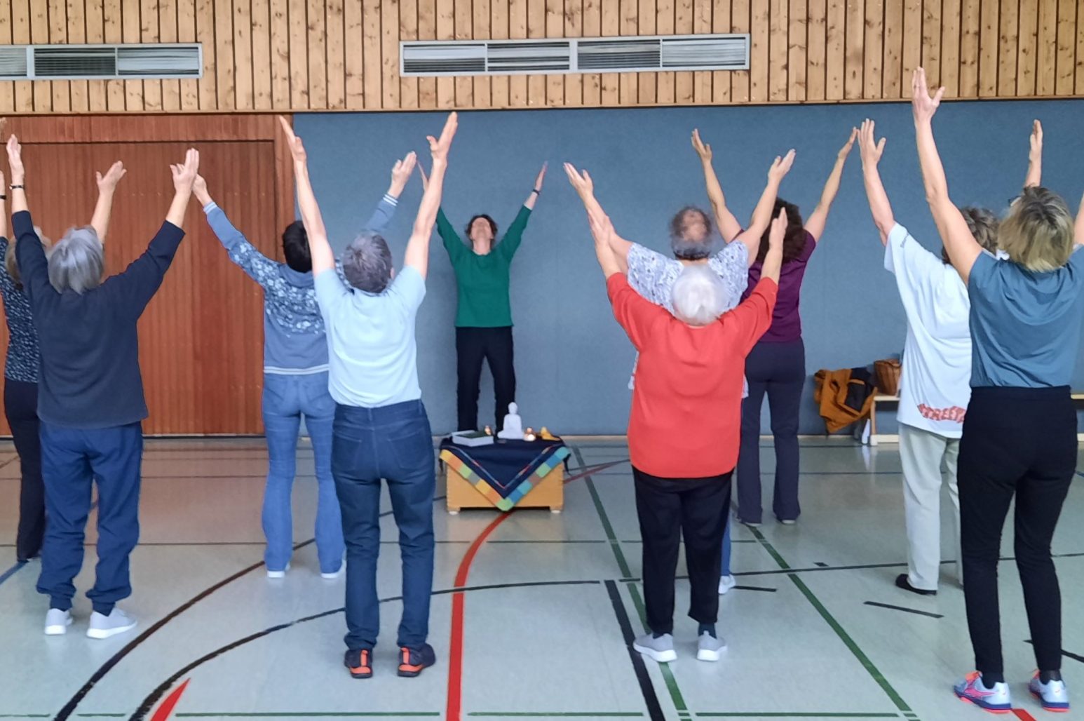 Eine Gruppe von Menschen steht mit erhobenen Händen in einer Sporthalle und machen Qigong.