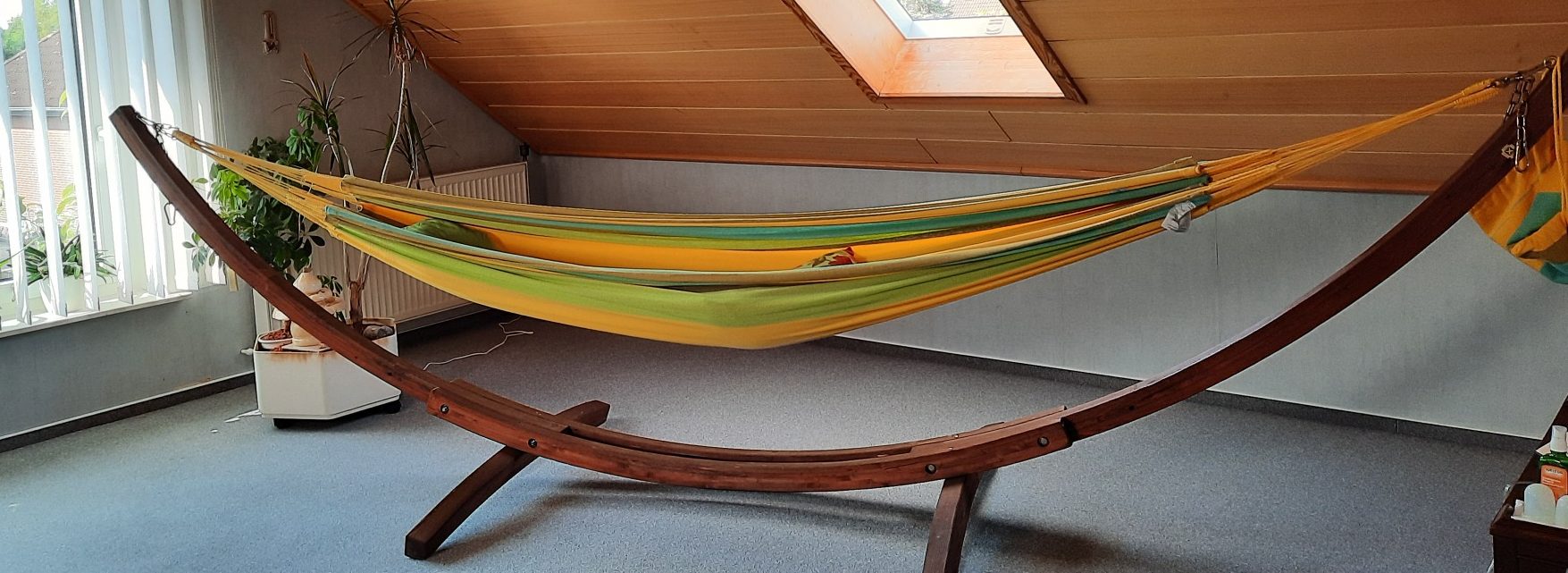 Hängematte in einem hellen Raum, auf einem Holzgestell, mit gelb-grüner Bespannung.